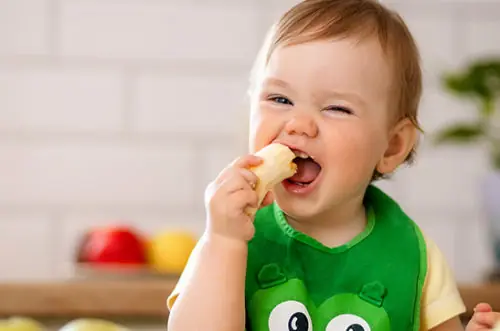 Bebê sorrindo enquanto come um pedaço de banana com as mãos