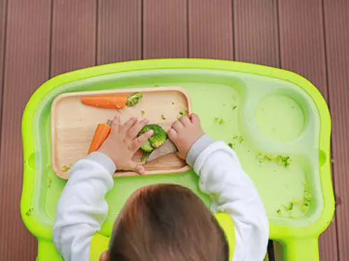 Bebê comendo brócolis e cenoura com as mãos no método BLW, visto de cima
