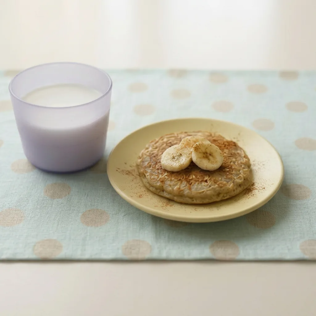 Panqueca de banana com aveia e canela em prato bege ao lado de copo de leite para bebê de 1 ano